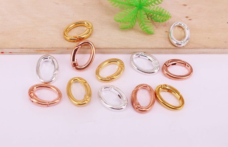 May include: An assortment of oval-shaped metal clasps in gold, silver, and rose gold. The clasps are open, showcasing their hinged design. The background is a light pink surface with a wooden element and a green plastic plant.