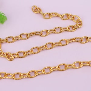 Op de afbeelding: Een gouden ketting met een getextureerde, ovale schakel. De ketting ligt op een roze oppervlak.