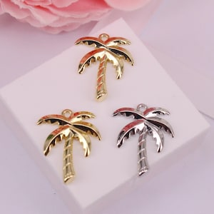 Op de afbeelding: Drie metalen palmboombedels in goud- en zilverkleur. De bedels hebben gedetailleerde bladeren en getextureerde stammen. Ze worden gepresenteerd op een wit oppervlak, met een zachte roze bloemenachtergrond.