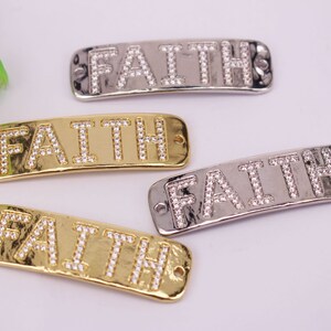 Puede incluir: Cuatro adornos metálicos rectangulares con la palabra "FAITH" escrita con brillantes pedrería. Dos son dorados y dos plateados. Cada uno tiene dos agujeros para sujetar.