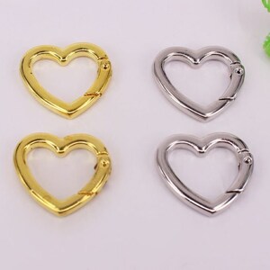 May include: Four heart-shaped metal clasps, two gold and two silver. The clasps are open and ready to be attached to a keychain, bag, or other item.