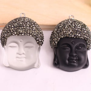 May include: Two Buddha head pendants with a silver tone metal loop. The pendants are made of white and black resin and are covered in black rhinestones.