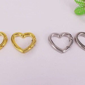 May include: Four heart-shaped metal clasps, two gold and two silver. The clasps are open and ready to be attached to a keychain, bag, or other item.