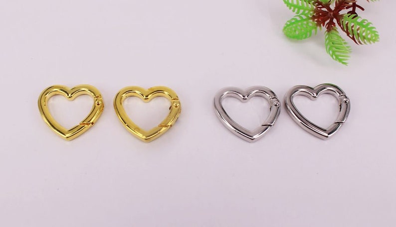 May include: Four heart-shaped metal clasps, two gold and two silver. The clasps are open and ready to be attached to a keychain, bag, or other item.