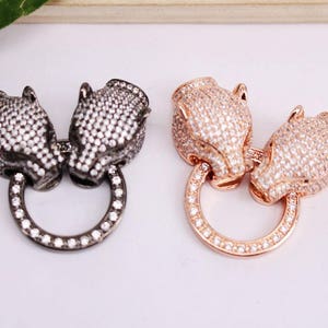 Könnte beinhalten: Zwei Tierkopf-Anhänger in Silber- und Goldtönen mit einem Kreisring. Die Anhänger sind mit klaren Strasssteinen besetzt.