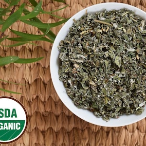 May include: A white bowl filled with dried, green herbs, possibly for tea or culinary use. The bowl sits on a woven, brown surface, with green leaves in the upper left corner. A USDA ORGANIC seal is visible.