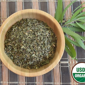 May include: A wooden bowl filled with dried, green, organic mint leaves. The bowl is sitting on a brown woven mat. A green USDA Organic seal is visible in the lower right corner of the image.