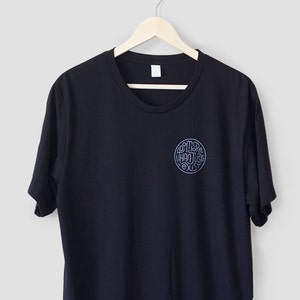 May include: A navy blue t-shirt with a round, light blue graphic on the upper left chest. The graphic features stylized text within a circular design. The t-shirt is hanging on a wooden hanger against a light gray background.