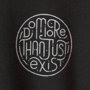 May include: Black fabric with a circular design in light gray. The design features the words "Do More Than Just Exist" in a stylized, hand-drawn font, enclosed within a dotted circle.