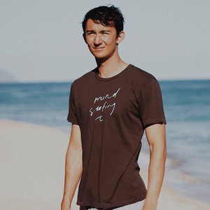 May include: A man wearing a brown t-shirt with the text "mind surfing" written in white. He is standing on a sandy beach with the ocean in the background.