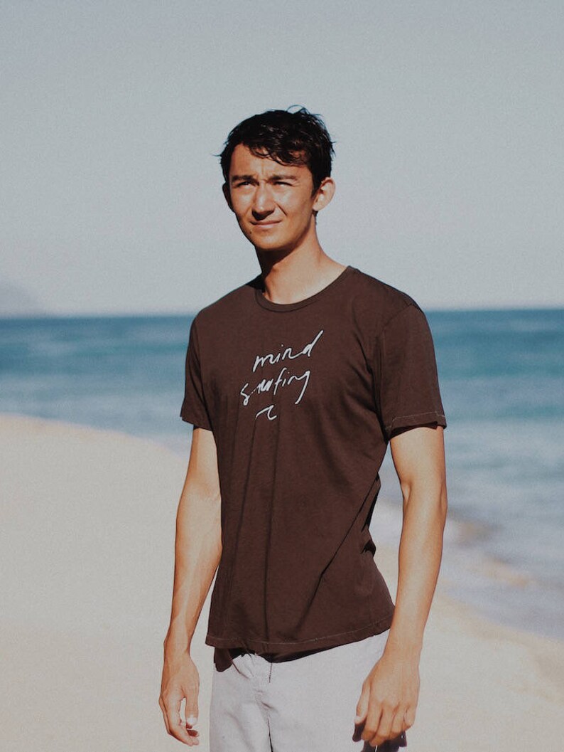 May include: A man wearing a brown t-shirt with the text "mind surfing" written in white. He is standing on a sandy beach with the ocean in the background.