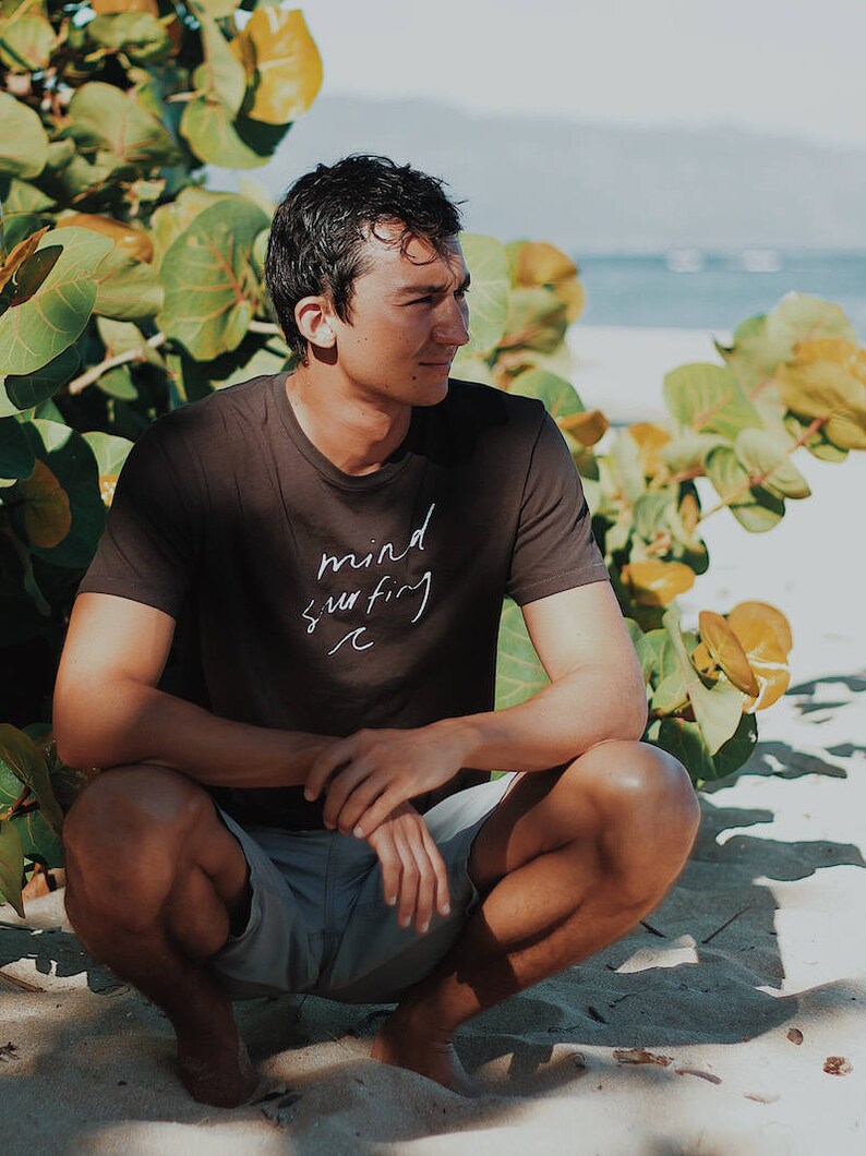 May include: A person wearing a black t-shirt with the text "mind surfing" in white lettering. The person is sitting on the sand with their legs crossed and looking to the right. The person is surrounded by green foliage.