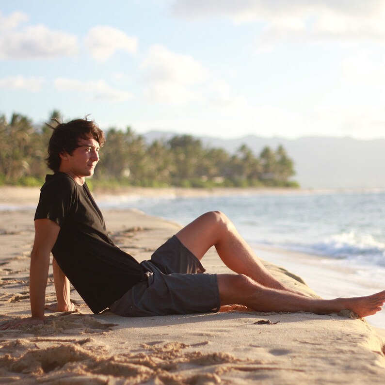 May include: A person wearing a black t-shirt and gray shorts is relaxing on a sandy beach. The person is sitting near the water's edge, with a backdrop of palm trees, ocean, and a partly cloudy sky.