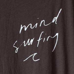 May include: A dark brown t-shirt with the words "mind surfing" written in white cursive script. A small wave graphic is below the text.