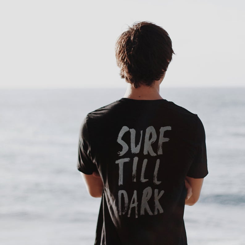 May include: A person wearing a black t-shirt with the words "Surf Till Dark" written in white on the back. The person is standing in front of a body of water.