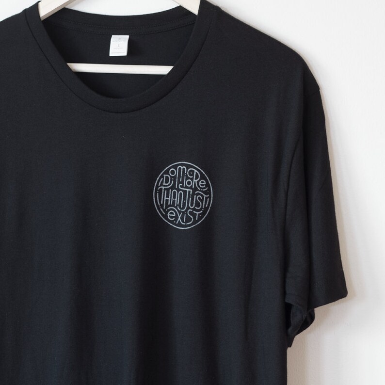 May include: Black crew neck t-shirt with a circular graphic on the chest. The graphic features the words "Do More Than Just Exist" in a stylized, handwritten font. The shirt is hanging on a white wooden hanger.