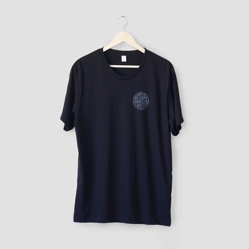 May include: A navy blue t-shirt with a round, light blue graphic on the upper left chest. The graphic features stylized text within a circular design. The t-shirt is hanging on a wooden hanger against a light gray background.