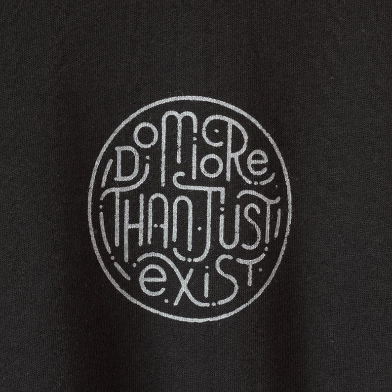 May include: Black fabric with a circular design in light gray. The design features the words "Do More Than Just Exist" in a stylized, hand-drawn font, enclosed within a dotted circle.