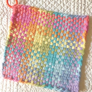 May include: A square potholder made of woven fabric featuring a multicolored grid pattern in shades of pink, orange, yellow, green, and blue. The potholder has a small, solid orange plastic ring attached to one corner for hanging purposes.