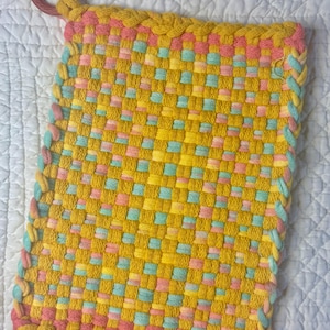 May include: A rectangular woven potholder featuring a vibrant yellow, pink, and light blue checkered pattern. The edges are finished with a braided border in matching colors, and a circular red plastic ring is attached to the top corner.