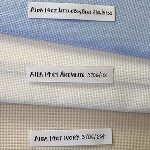 May include: Three Aida fabric swatches in different colors: light blue, white, and ivory. Each swatch has a label with the text "AIDA 14CT" and the color name and corresponding product code.