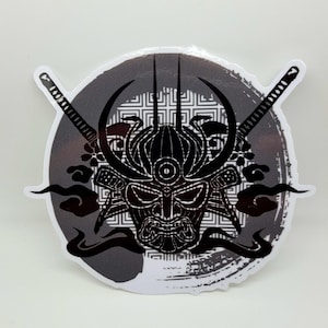 May include: Black and white sticker featuring a samurai warrior mask with a third eye, surrounded by stylised smoke and crossed swords. The background is a grey circle with a geometric pattern.