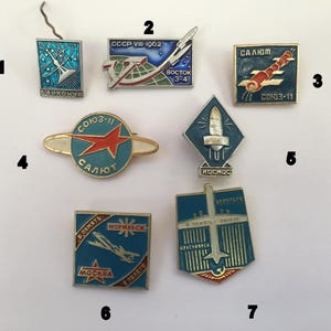 May include: A collection of vintage space-themed pins. The pins feature rockets, satellites, and text in Cyrillic script. Colors include blue, red, gold, and silver. The pins are likely from the Soviet era and commemorate space exploration.