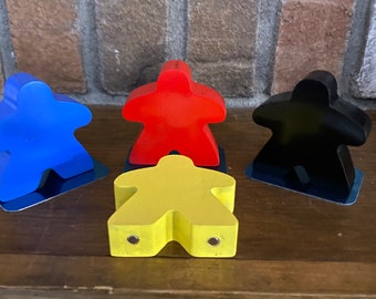 May include: Four meeple game pieces in different colors: blue, red, black, and yellow. The meeples are standing on a wooden surface.