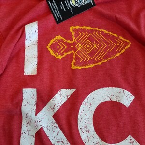 May include: Red hoodie with the text "I KC" in distressed white lettering. A yellow arrow graphic is above the "KC". A tag with a skull design and text is attached to the hoodie.