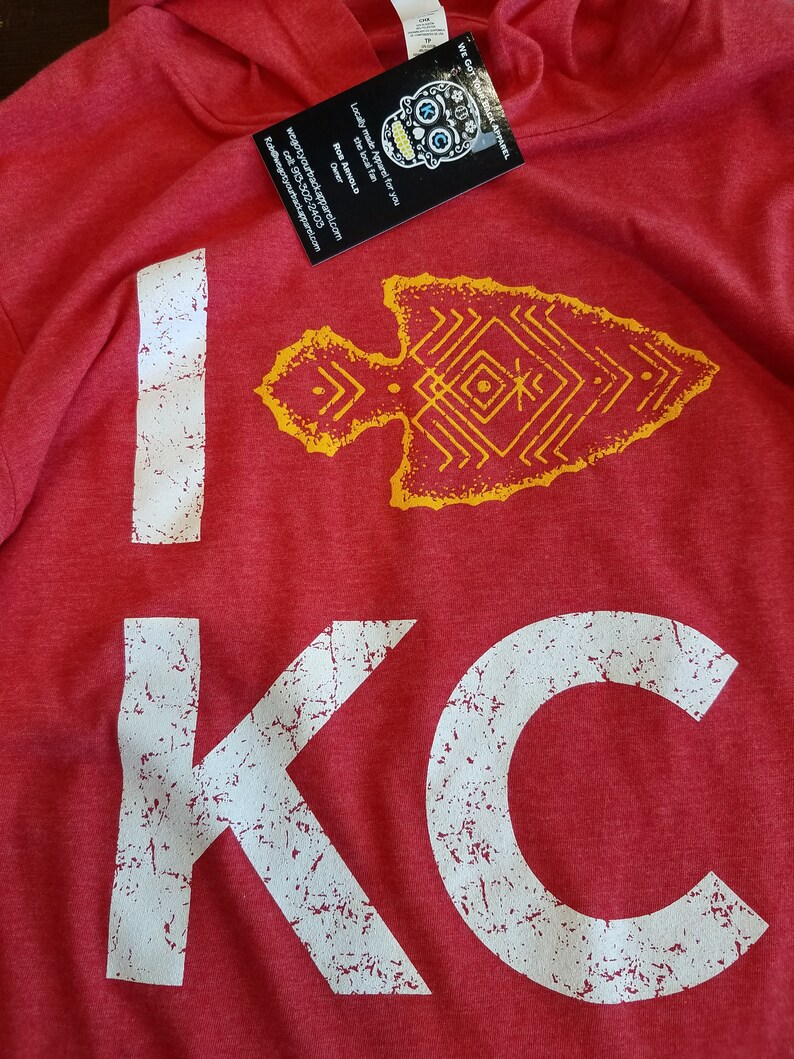 May include: Red hoodie with the text "I KC" in distressed white lettering. A yellow arrow graphic is above the "KC". A tag with a skull design and text is attached to the hoodie.