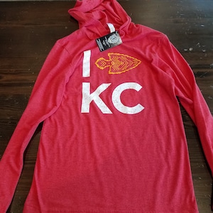 May include: Red long-sleeve hooded shirt with the text "I KC" in white distressed font. A yellow and orange arrowhead graphic is above the "KC". The shirt is lying on a dark wood surface.