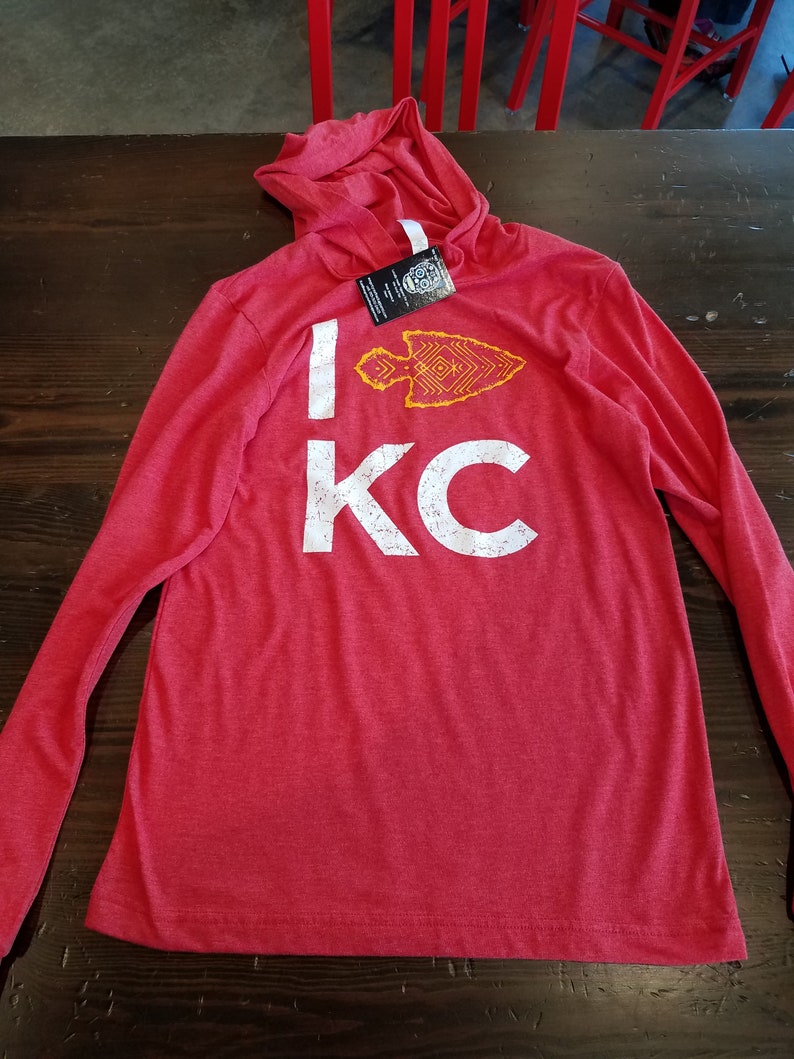 May include: Red long-sleeve hooded shirt with the text "I KC" in white distressed font. A yellow and orange arrowhead graphic is above the "KC". The shirt is lying on a dark wood surface.