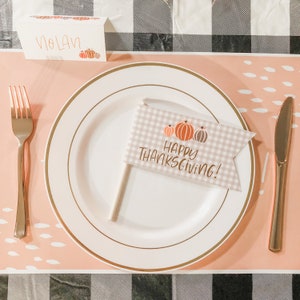 May include: Thanksgiving table setting with a white plate, gold rim, and a flag that says "Happy Thanksgiving!" with pumpkin illustrations. A fork and knife are on either side of the plate. A place card with the name "Nolan" is at the top.