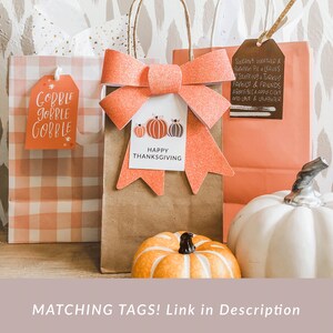May include: Thanksgiving-themed gift bags with orange glitter bows and tags. One tag reads "Gobble Gobble Gobble," another says "Happy Thanksgiving," and a third lists autumn-related phrases. A decorative pumpkin and a white pumpkin are also visible.