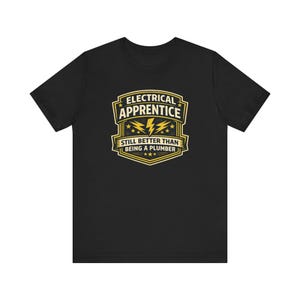 Puede incluir: Camiseta negra con un gráfico dorado y blanco que dice "ELECTRICAL APPRENTICE" sobre un diseño de rayo. El texto "STILL BETTER THAN BEING A PLUMBER" está debajo del gráfico. La camiseta está hecha de un material suave.
