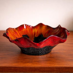 May include: A decorative ceramic bowl with a ruffled edge. The bowl features a vibrant color scheme, with a red rim that transitions to orange and yellow in the center. The base is a textured black.