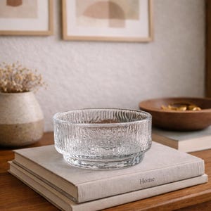 May include: A clear glass bowl with a textured, ridged design sits on a stack of books. The bowl is round with a wide base and a textured exterior. The word "Home" is visible on the book. The background includes a wooden bowl and framed artwork.
