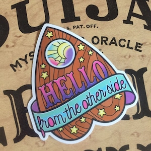 May include: A brown wooden Ouija board shaped like a heart with a blue, pink, and purple gradient. The board has a crescent moon and stars on it. The text "HELLO from the other side" is written in a white banner.