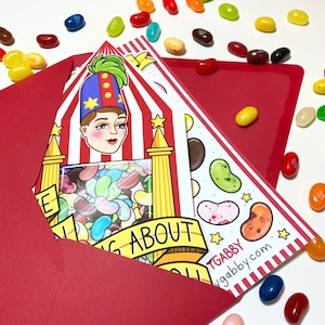 May include: A red envelope with a white and red striped card inside. The card features a cartoon illustration of a person with a red and blue hat, a red and white striped shirt, and a red and white striped background. The card also features a clear plastic window filled with colorful jelly beans. The text on the card reads "Thinking about you". The card is signed "TGABBY" and includes the website "tgabby.com".
