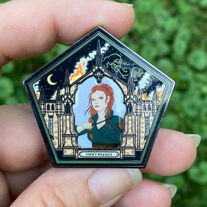 May include: A black and gold enamel pin shaped like a Hogwarts house crest. The crest features a red-haired woman holding a wand in front of a burning building. The text "Ginny Weasley" is at the bottom of the pin.