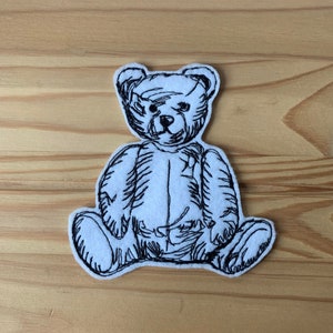 May include: Black and white embroidered patch of a teddy bear sitting down. The bear is outlined in black thread and has a simple, cartoon-like design.