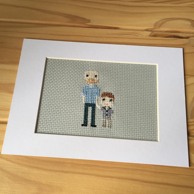 May include: A framed cross-stitch artwork depicting two pixelated figures, a father and son, standing side-by-side. The father has brown hair and a beard, wearing a blue shirt and dark pants. The son has brown hair and is wearing a light blue shirt and dark pants.
