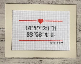 Pode incluir: Um bordado de ponto de cruz enquadrado em branco com fio vermelho e preto. O bordado apresenta um coração vermelho, as coordenadas "34°59'24"N 33°58'41"E" e a data "6-6-2017".