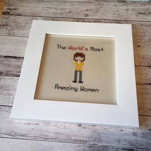 Op de afbeelding: Een wit ingelijst kruissteek met een gepixelde afbeelding van een persoon met bruin haar, die een geel shirt, een grijze broek en zwarte schoenen draagt. De tekst "The World's Most Amazing Woman" is in rood en zwart geborduurd.