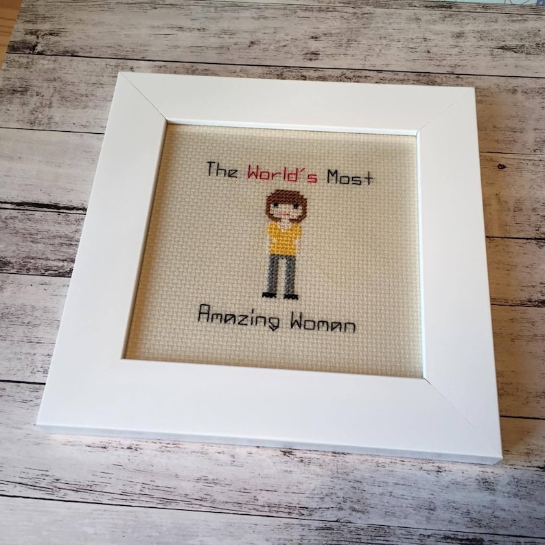 Op de afbeelding: Een wit ingelijst kruissteek met een gepixelde afbeelding van een persoon met bruin haar, die een geel shirt, een grijze broek en zwarte schoenen draagt. De tekst "The World's Most Amazing Woman" is in rood en zwart geborduurd.