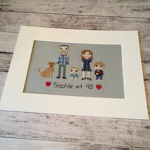 Puede incluir: Bordado de punto de cruz de una familia con un perro. La familia incluye dos adultos, un ni&ntilde;o y un beb&eacute;. El bordado est&aacute; enmarcado e incluye el texto "Sophie at 40".