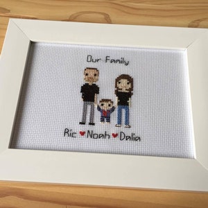 May include: A white framed cross-stitch with a pixelated image of a family of three. The text "Our Family" is above the image and "Ric Noah Dalila" is below the image.