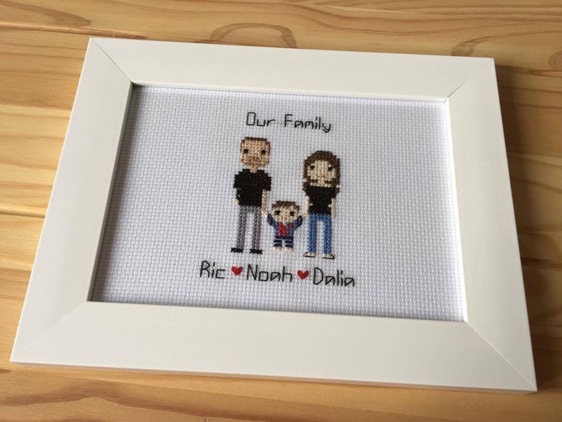 May include: A white framed cross-stitch with a pixelated image of a family of three. The text "Our Family" is above the image and "Ric Noah Dalila" is below the image.