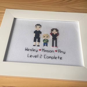 Op de afbeelding: Witte ingelijste kruissteek met een familieportret van drie personen. De kruissteek toont een man, een vrouw en een kind. De tekst onder de afbeelding luidt "Wesley &hearts; Mason &hearts; Amy Level 2 Complete".