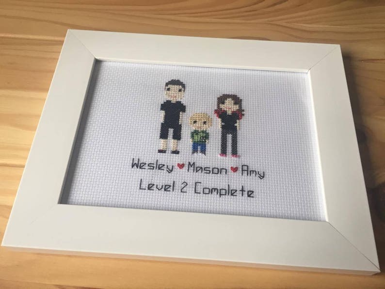 Op de afbeelding: Witte ingelijste kruissteek met een familieportret van drie personen. De kruissteek toont een man, een vrouw en een kind. De tekst onder de afbeelding luidt "Wesley &hearts; Mason &hearts; Amy Level 2 Complete".